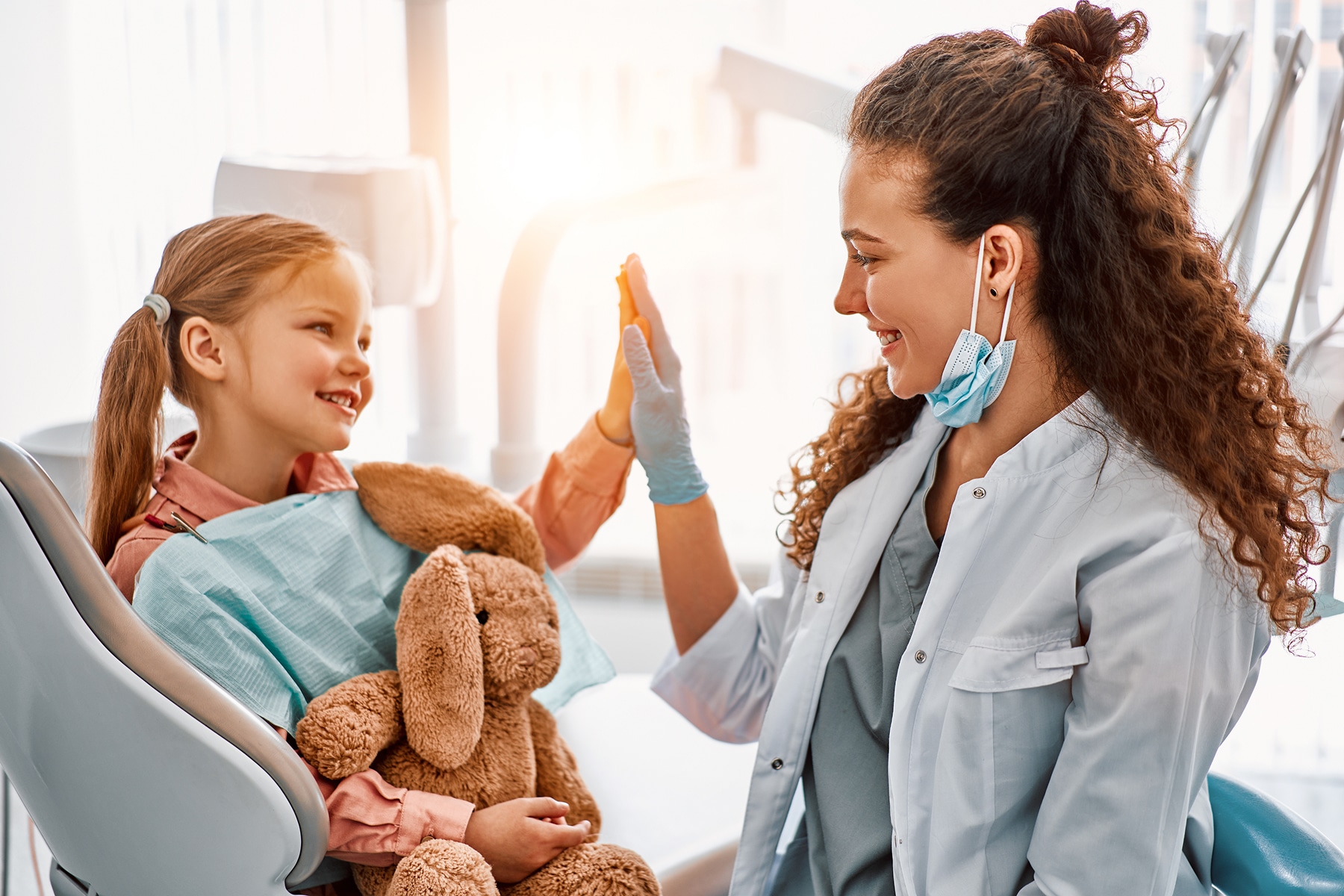 A child with a stuffed toy in a dental chair gives a high-five to a person wearing a mask and gloves in a dental clinic.