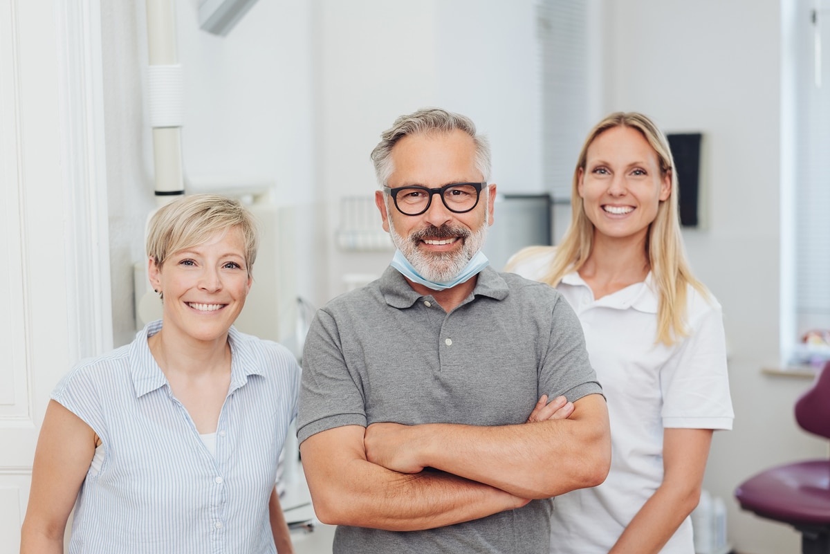 Three smiling people in a dental office setting; one wears dental attire with a mask around the neck, suggesting a professional environment.
