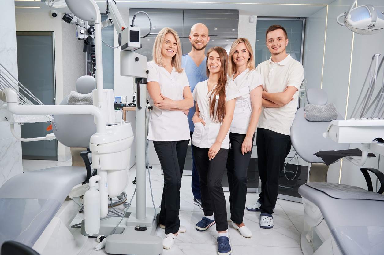 Five people in a modern dental clinic, smiling, standing among dental equipment and chairs, wearing coordinated outfits in a clinical setting.