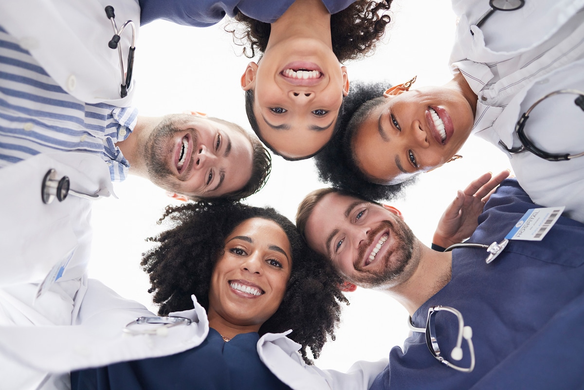 Five smiling healthcare professionals form a circle, looking down into the camera. They wear medical attire with stethoscopes, symbolizing teamwork and unity.