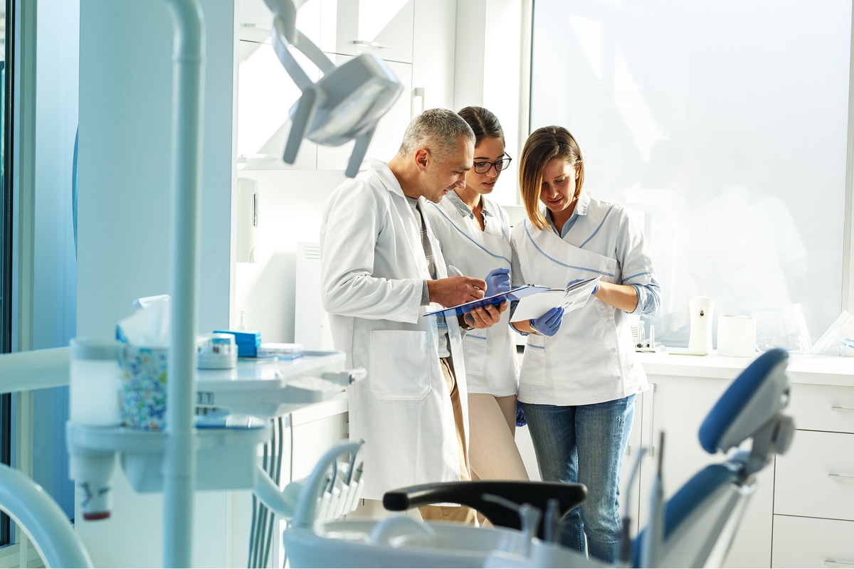 Three people in lab coats review documents in a bright dental office, surrounded by dental equipment and tools.