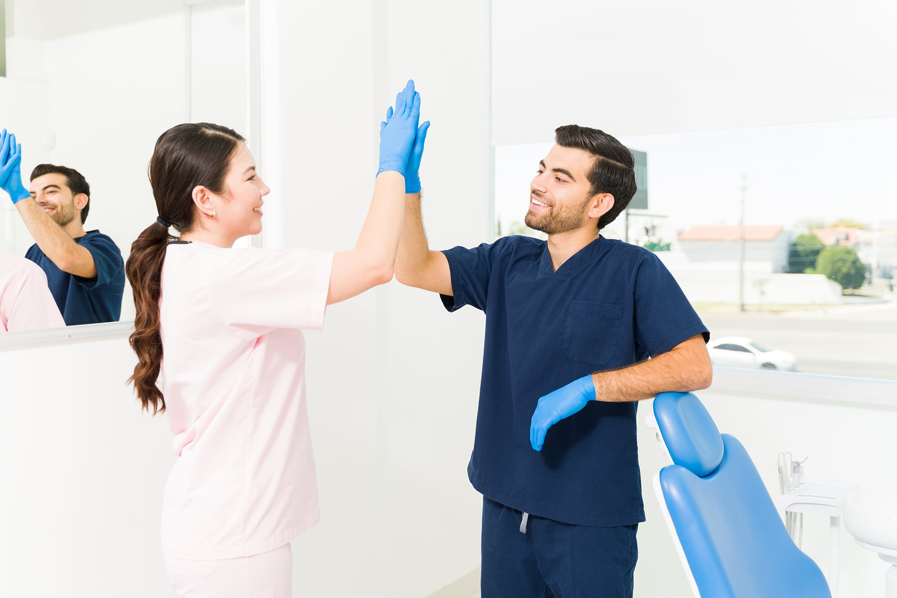 Two people in scrubs and gloves high-five in a dental office. A dental chair and reflection in the mirror are visible.
