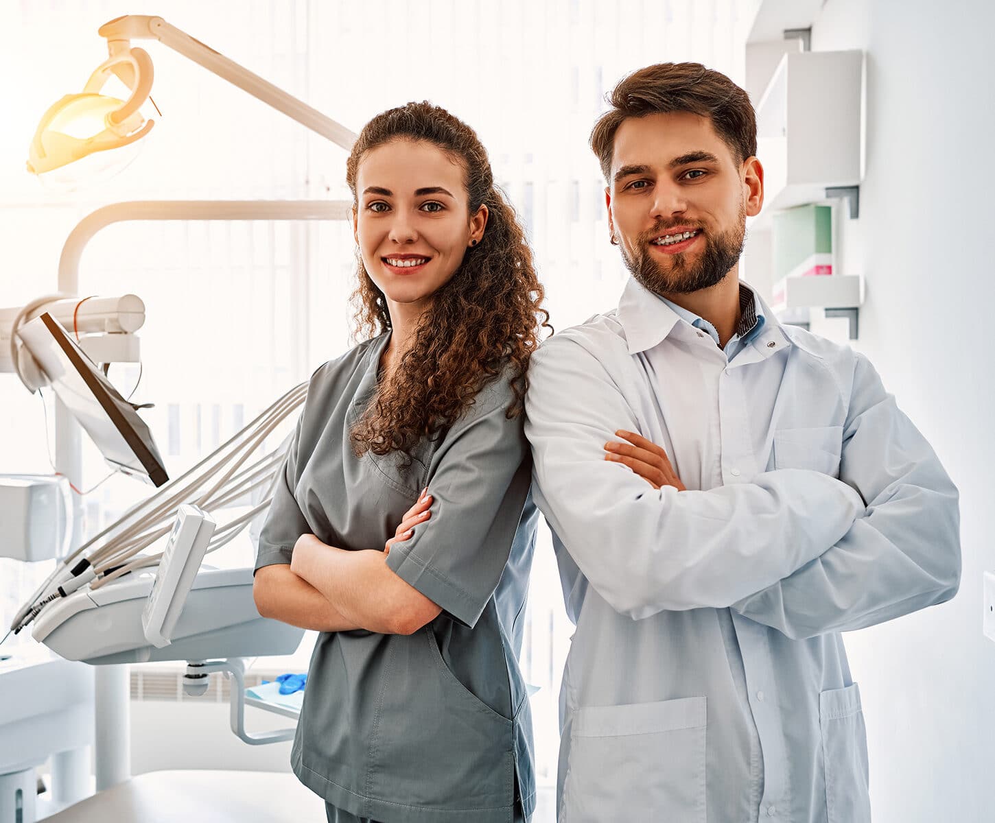 Two people in dental attire stand in a modern dental office, smiling confidently, with dental equipment visible in the background.