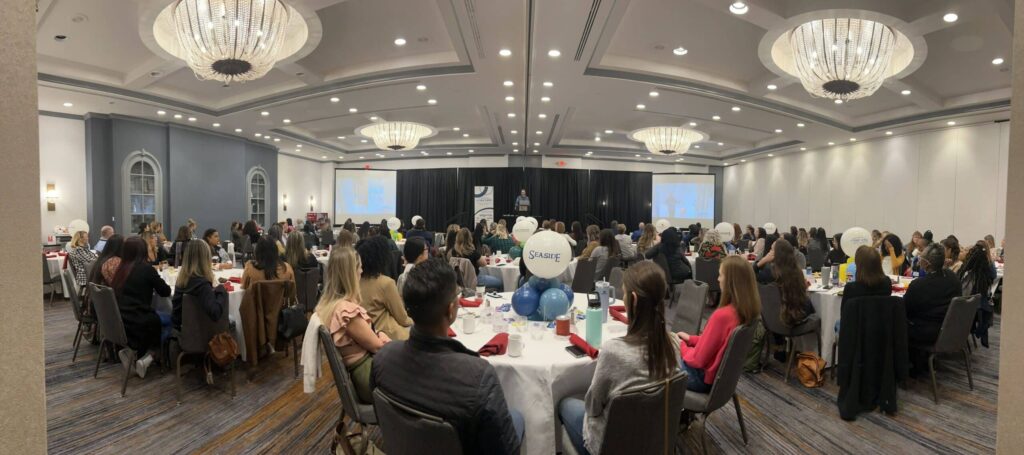 A conference room with chandeliers hosts round tables of people attending a presentation. Balloons and a speaker are visible on stage.